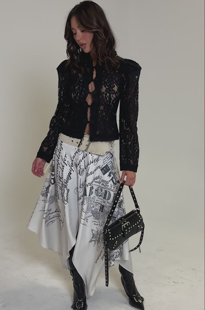 Video of woman modeling the READ ME MIDI SKIRT with black newspaper-style graphics, black knee-high boots, and holding a black studded handbag.