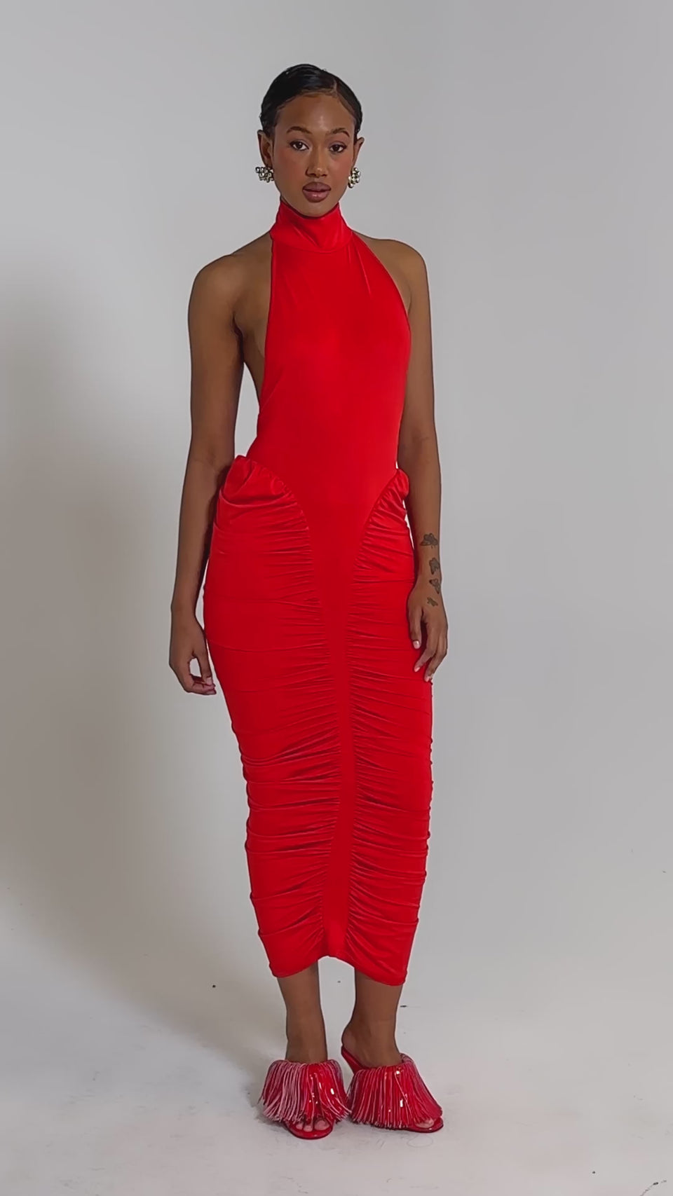 A video of a person in the HIPS DON'T LIE RUCHED MAXI DRESS IN RED, a sleeveless, high-neck, fitted dress with ruching, styled with varsity-inspired red shoes featuring fringe accents against a plain light background.