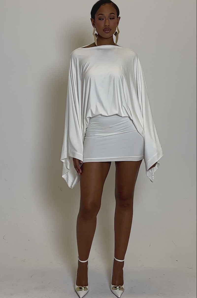 Video of woman modeling the DIANA DRAPED MINI DRESS IN WHITE with dramatic batwing sleeves and a fitted skirt, styled with large geometric gold earrings.