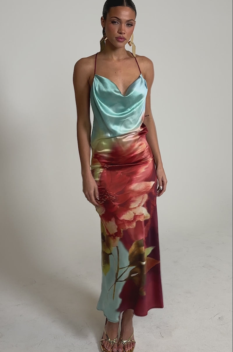 Video of woman modeling the WORK OF ART SATIN MAXI DRESS, a sleeveless maxi with a blue top blending into a red skirt adorned with large floral prints. She pairs it with gold high-heeled sandals, statement earrings, and has slicked-back hair.