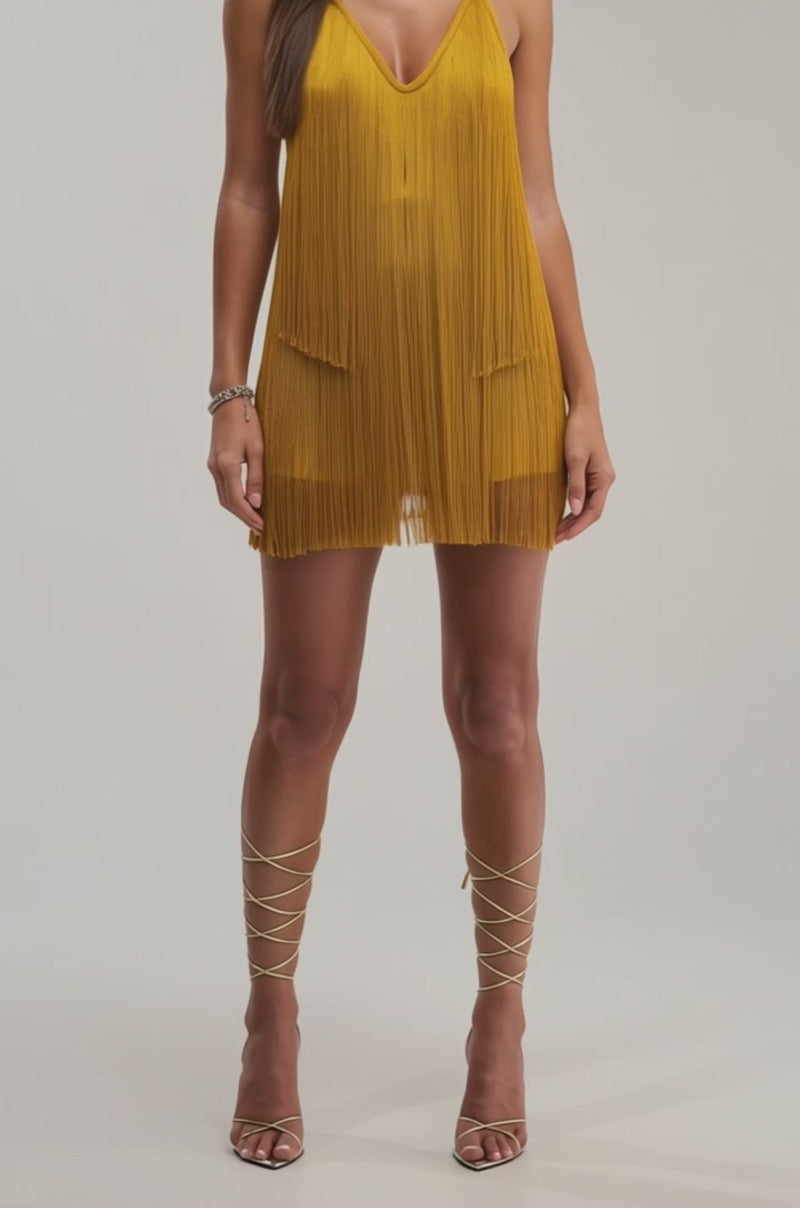A video of a woman wearing the DRINKS ON ME FRINGE MINI DRESS IN GOLD, featuring a V-neckline and fringe, standing against a plain light background.
