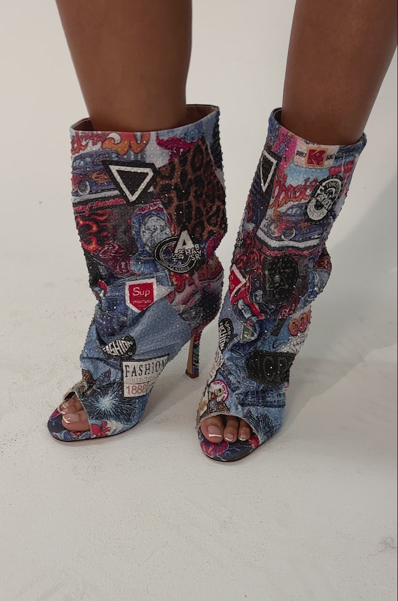 Video of woman modeling the AZALEA WANG JONICE BLUE MULTI RHINESTONE OPEN TOE BOOTIE, featuring colorful patterns and fashion-themed patches, while standing against a plain background.
