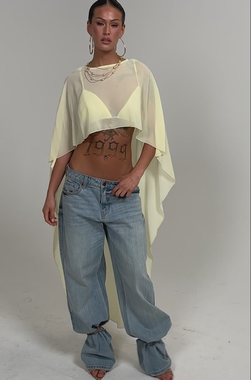 Video of woman modeling the COOL GIRL CHIFFON BLOUSE IN YELLOW, styled over a crop top with loose light blue jeans, open-toe heels, a woven handbag, large hoop earrings, and visible tattoos.