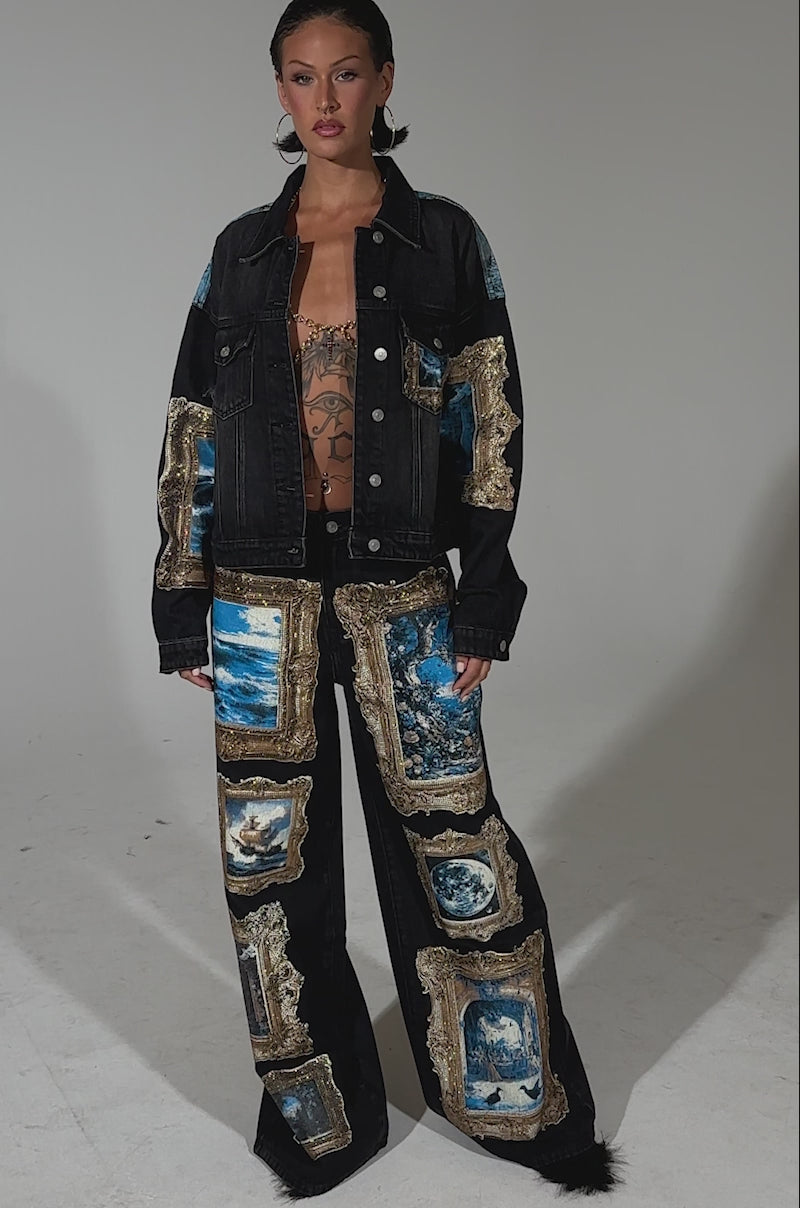 A video of a woman wearing the WORK OF ART GRAPHIC DENIM JACKET, featuring rhinestone trim and blue and gold framed artwork prints, styled with matching wide-leg pants, slicked-back hair, and a brown graphic crop top underneath.