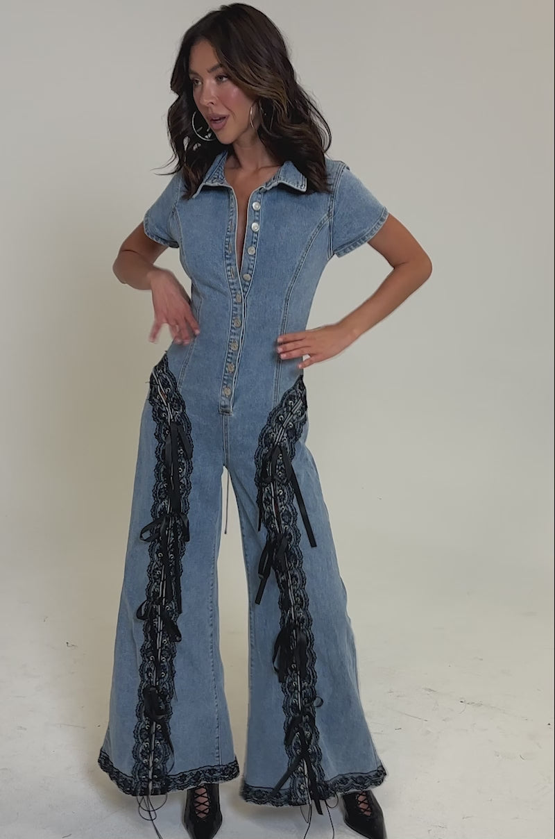 Video of woman modeling the DIXIE LACE DENIM JUMPSUIT, a short-sleeved, button-up piece with black lace sides and front, styled with black lace-up boots and loose wavy hair against a plain light background.