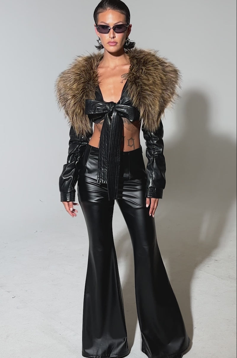 A video of a person modeling the SPEND IT ALL FUR TRIM BOLERO with black flared leather pants and a matching crop top, accessorized with sunglasses and hoop earrings, their hair slicked back, standing confidently against a plain background.
