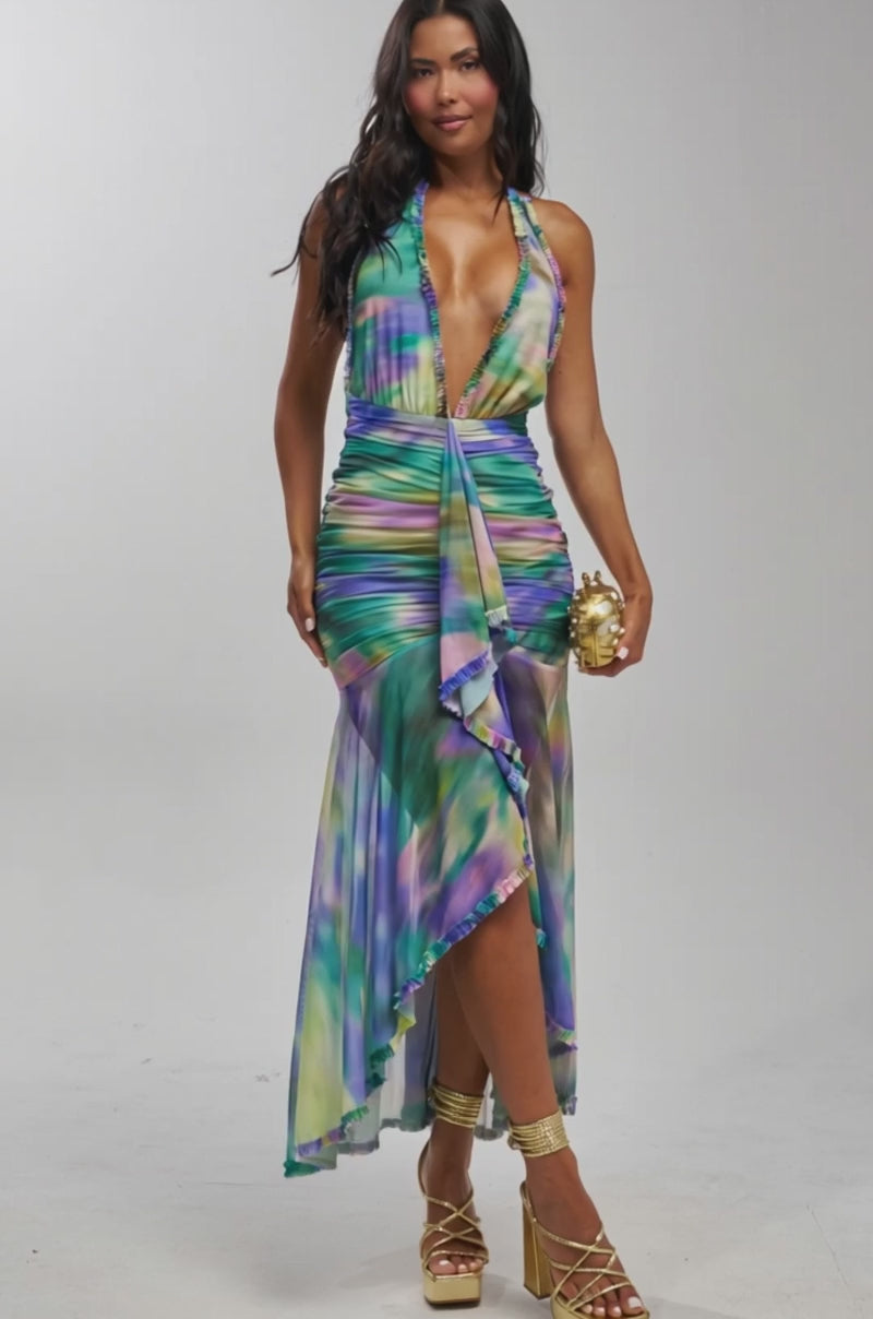 Video of woman modeling the CAMERA READY MAXI DRESS IN PURPLE MANIA, a sleeveless, deep V-neck mermaid dress in blue, green, and purple tie-dye with a ruched skirt and asymmetrical hem. She carries a gold clutch and wears gold platform heels.
