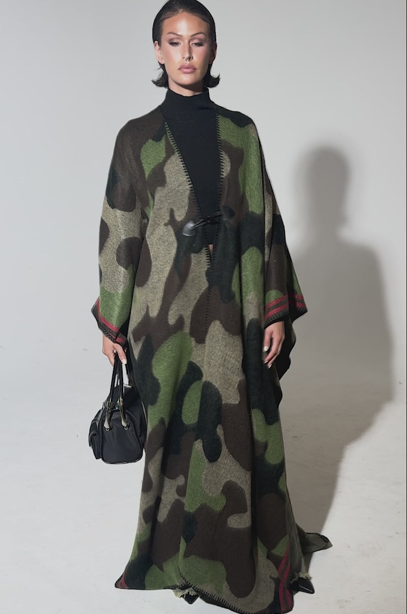 A video of a model wearing the CAMO NIGHTS MAXI PONCHO with red trim over a black turtleneck, a woman holds a black handbag and gazes confidently at the camera against a simple background.