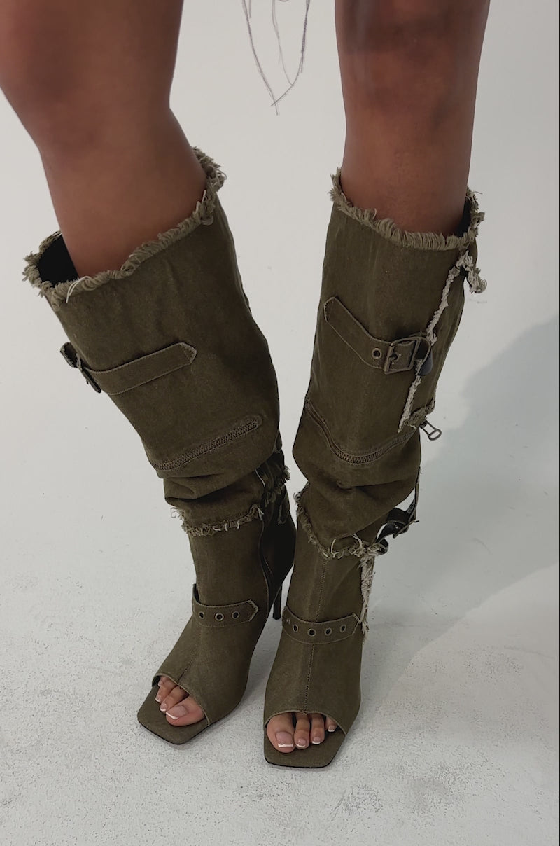 Video of woman modeling the AKIRA GOLD NADAL OLIVE CANVAS OPEN TOE STILETTO BOOTIES—knee-high, heeled with exposed seams, buckles, frayed edges, open toes, and side zippers.