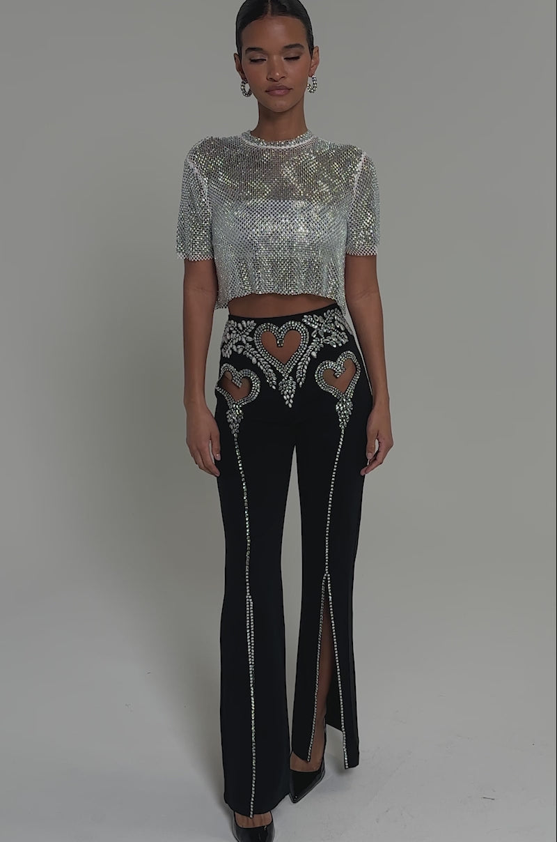 Video of woman modeling the LOVE FOR YOU RHINESTONE TROUSER—black flared pants with heart cutouts, crystal embellishments, front slits revealing high heels—paired with a sparkly cropped top, all set against a light background.