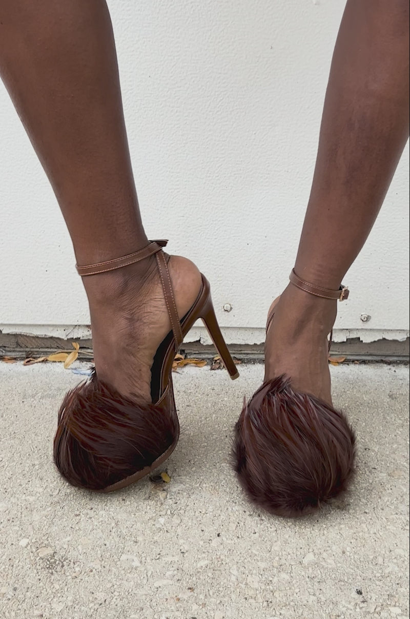 Video of woman modeling the AZALEA WANG ANGELES BROWN FEATHER STILETTO SANDAL, featuring brown feather accents and vegan leather, posing on a concrete surface against a white background.