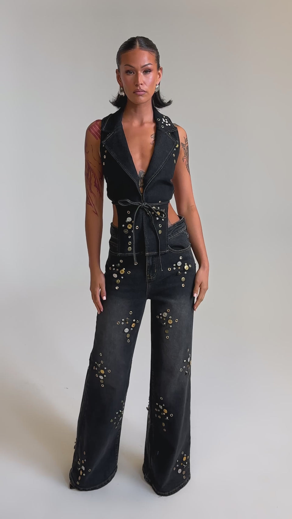 A video of a woman in the SPACE AND TIME STUDDED DENIM VEST—a dark-wash, sleeveless cropped top with gold accents—paired with matching wide-leg pants. Her slicked-back hair and confident pose, hand in pocket, complete the bold ensemble.