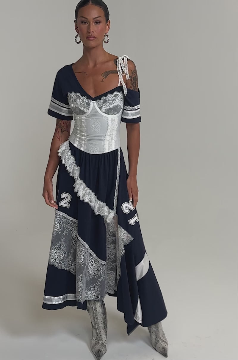 Video of woman modeling the BOX SUITE LACE JERSEY MAXI DRESS, featuring dark blue and white patchwork, lace accents, varsity numbers 12 and 33, an off-shoulder design, and white boots against a plain light backdrop.