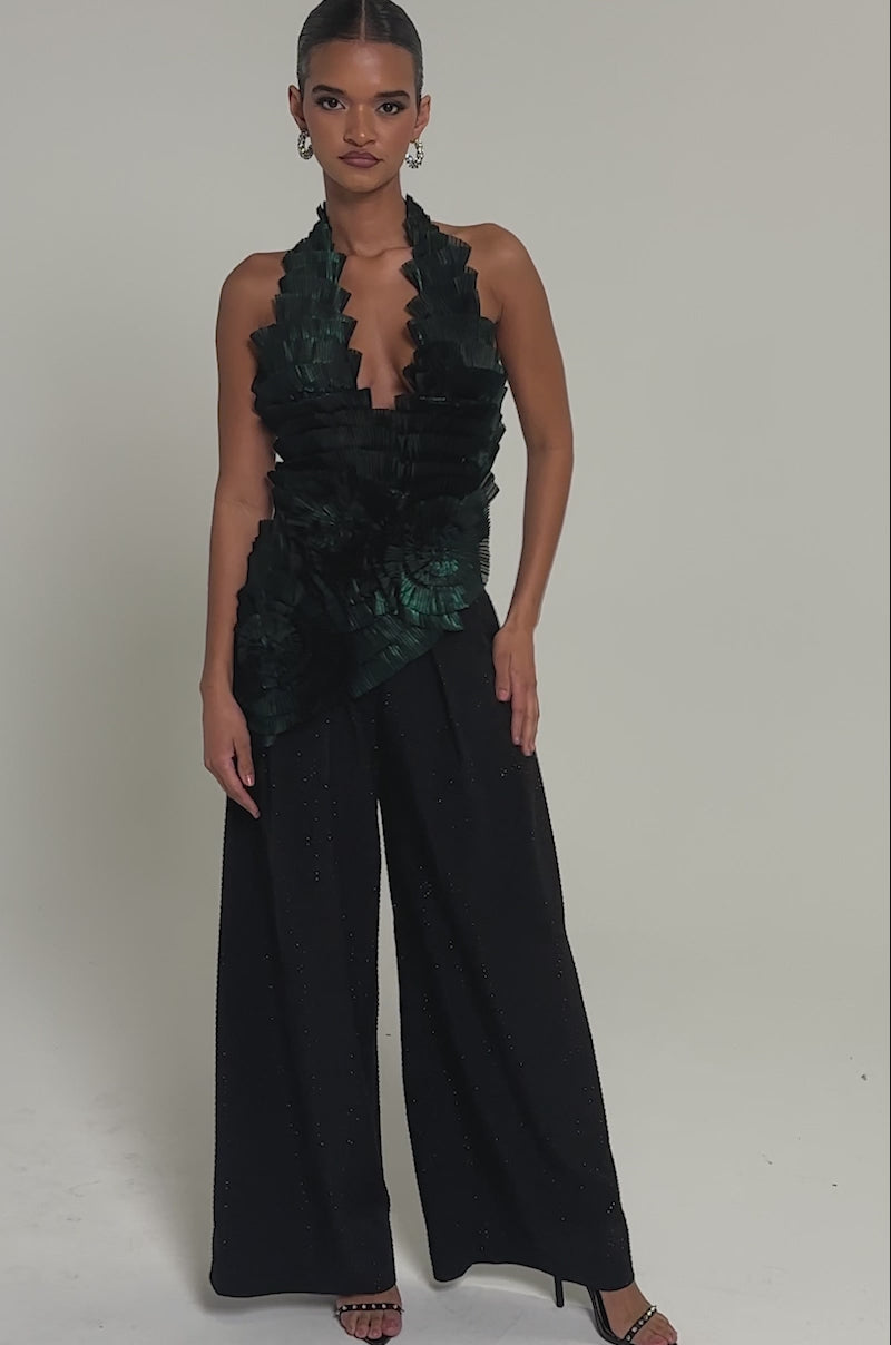 Video of woman modeling the DREAMERS HALTER BLOUSE in emerald green, showcasing layered fan-like pleats and circular floral motifs, paired with black pants against a neutral background.