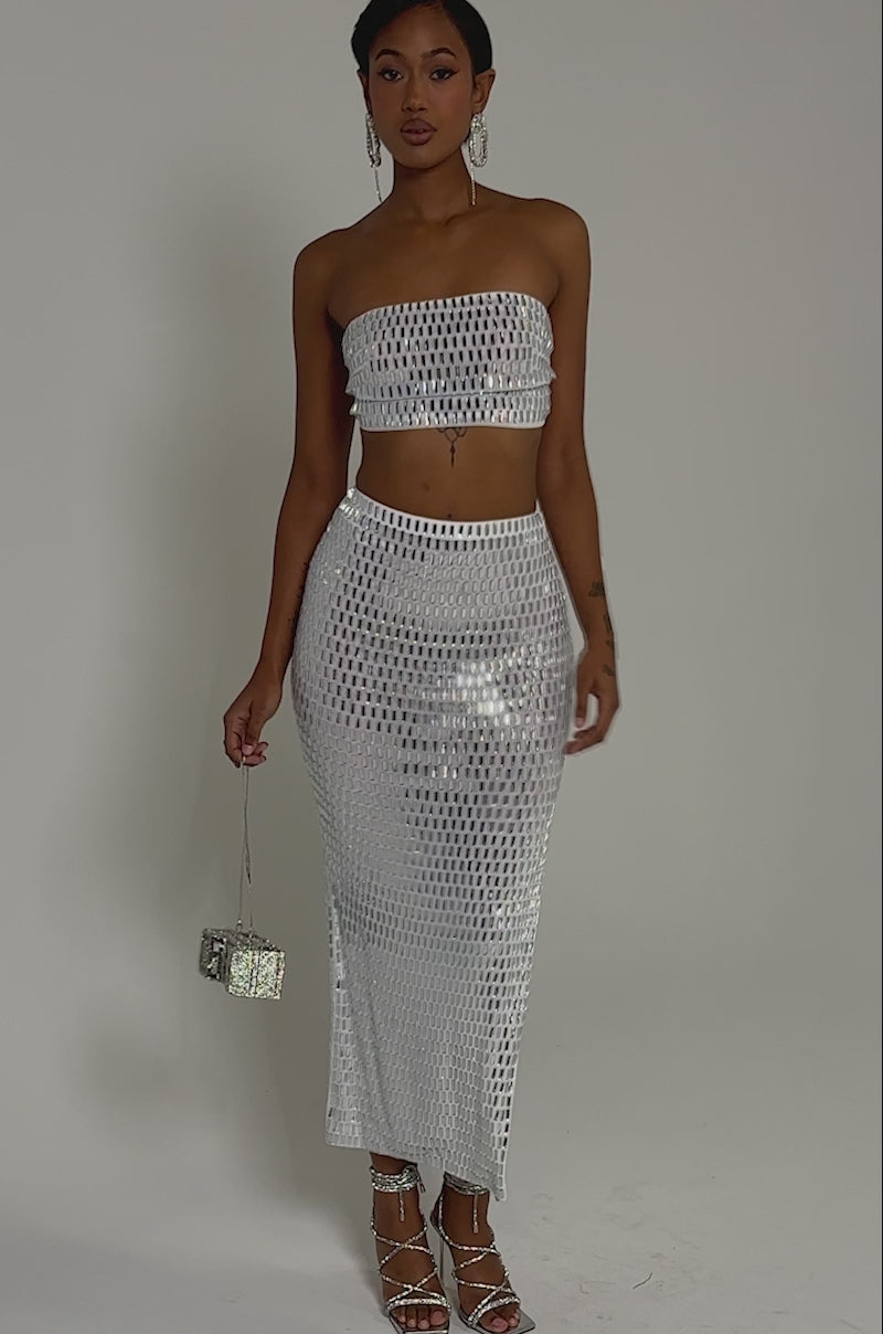 Video of woman modeling the AKIRA Label white, strapless crop top and the TALK ABOUT ME RHINESTONE MAXI SKIRT. She accessorizes with silver heels, statement earrings, and sleek straight hair.