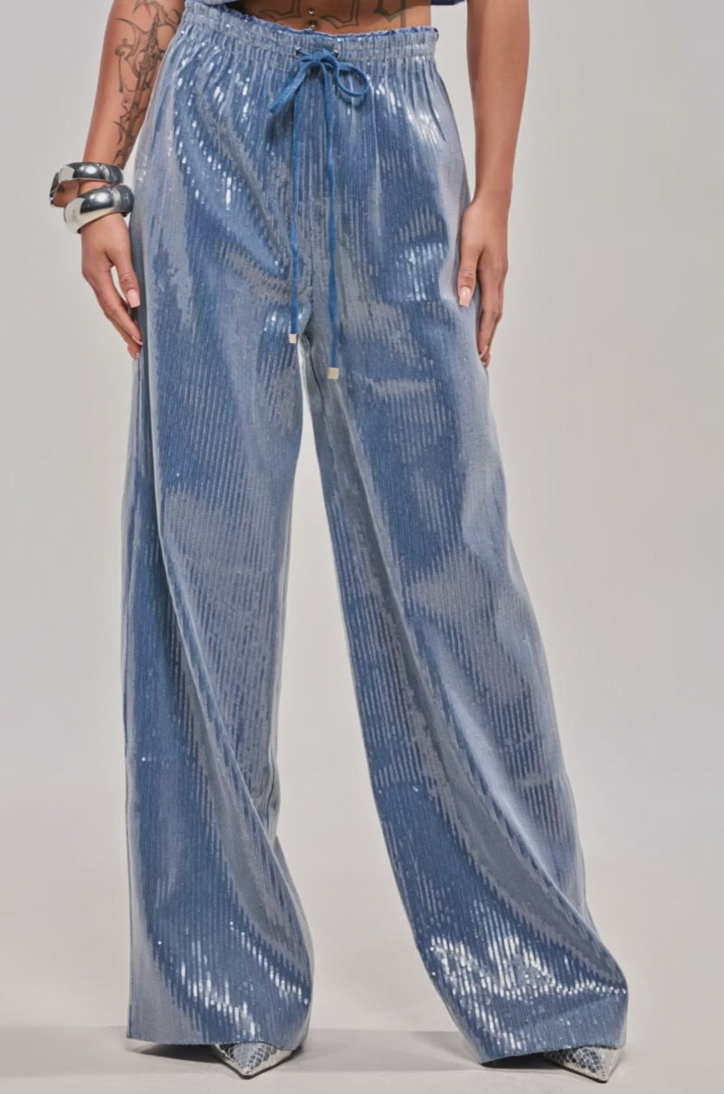Video of woman modeling the SHAKE IT GIRL SEQUIN WIDE LEG JOGGER in light blue, paired with a cropped short-sleeve button-up shirt and sparkling accessories, set against a plain light gray background.
