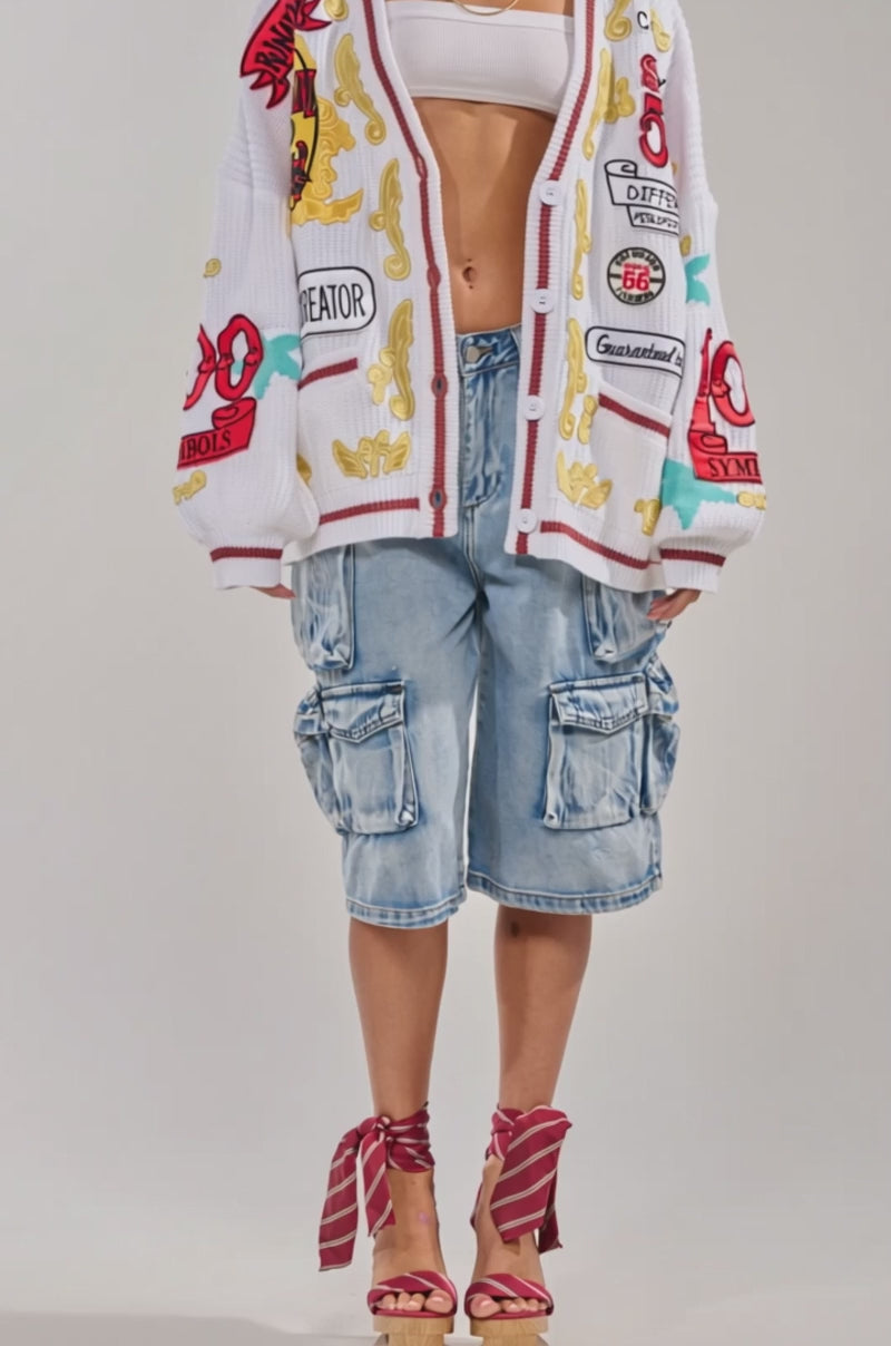 Video of woman modeling the NEW REGALIA PATCH CARDIGAN with bold red, gold, and blue patterns, large sunglasses, and light blue cargo jeans stands against a plain background. The oversized white knit cardigan features colorful patches and symbols.