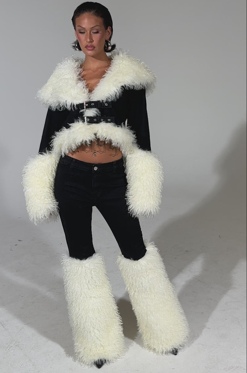 A video of a person wearing the SITTING PRETTY FUR TRIM DENIM set: a black crop top with fluffy white faux fur collar and cuffs, high-rise black jeans, and matching white faux fur leg warmers against a plain light gray background.