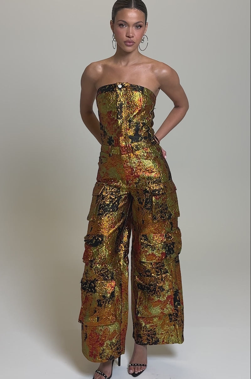 Video of woman modeling the NIKKI BROCADE JUMPSUIT, a strapless, wide-leg jumpsuit with cargo pockets and a bold orange, yellow, and black abstract print. She completes the look with hoop earrings and black heeled sandals.