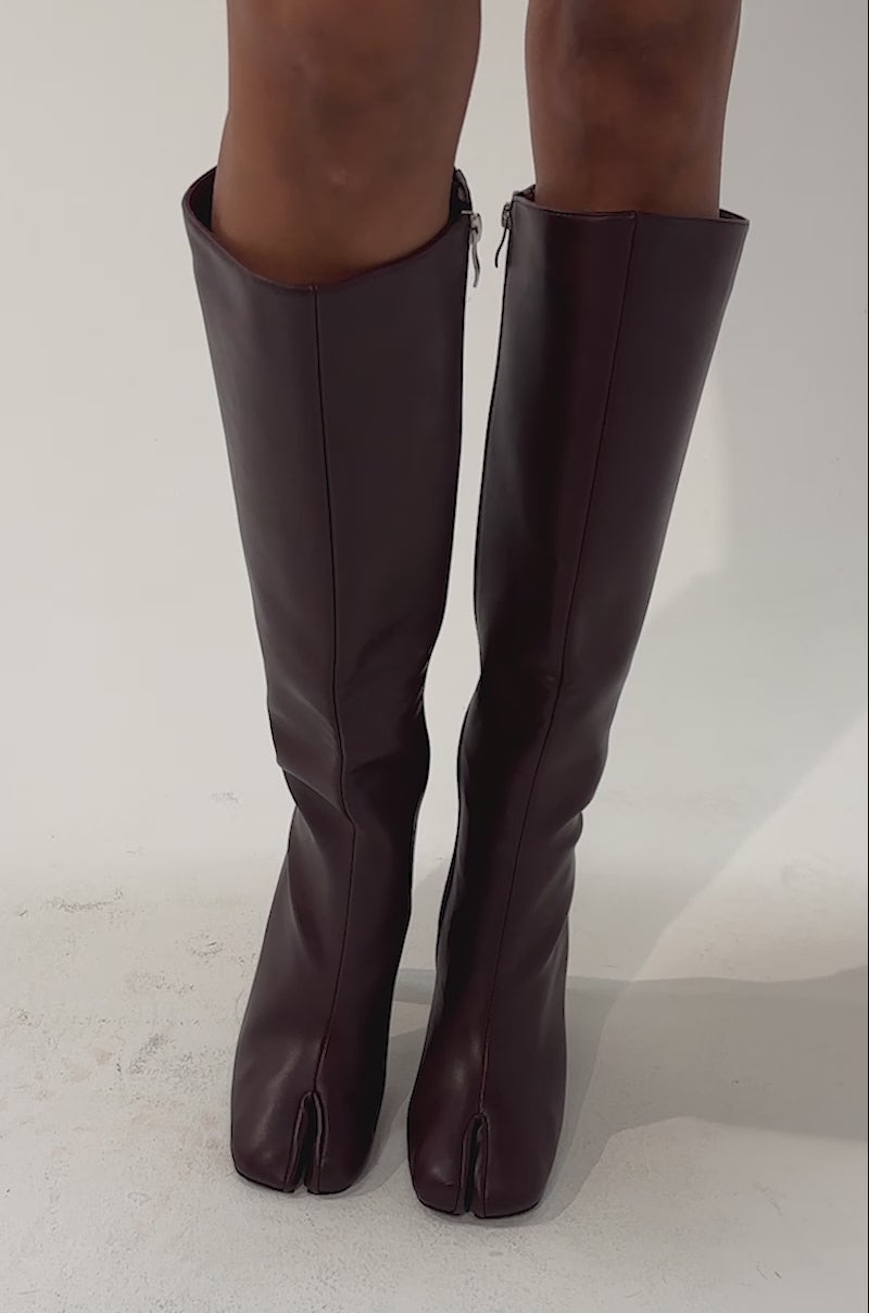 Video of a woman modeling the AZALEA WANG BILHANA BURGUNDY SPLIT TOE BLOCK BOOT—knee-high, burgundy boots crafted from leather, featuring a split-toe design and high block heels.