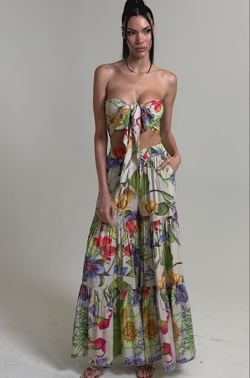 A video of a woman wearing the INTO THE GARDEN FLORAL BLOUSE, a vibrant tropical print halter-neck crop top, styled with a high-waisted tiered maxi skirt and sandals, standing confidently against a plain background.