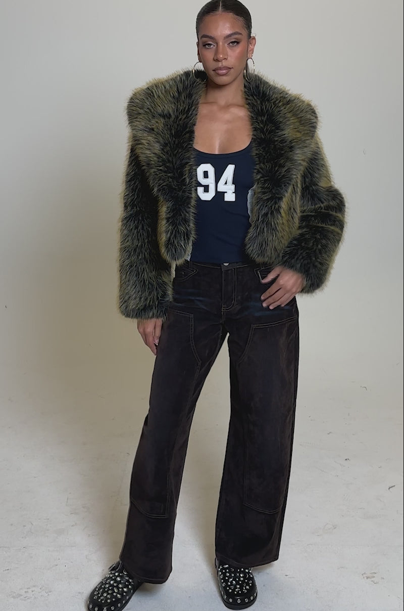 A video of a woman in the LIMONCELLO FAUX FUR COAT—yellow and black with an oversized shawl collar—paired with a navy "94" tank, brown wide-leg pants, black studded shoes, hoop earrings, and her hair pulled back.