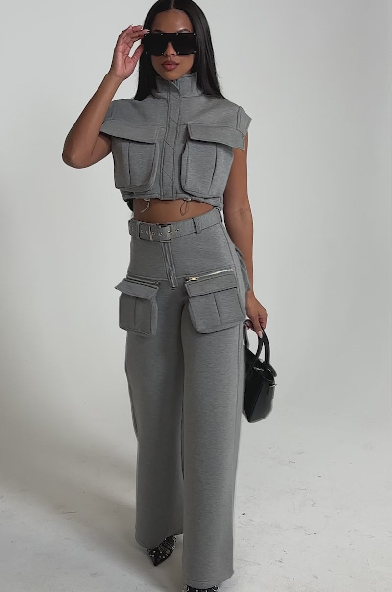 A video of a woman wearing the THAT GIRL SCUBA TOP, a sleeveless gray cargo blouse with large front pockets, paired with matching wide-leg cargo pants, oversized sunglasses, a black handbag, and studded shoes.