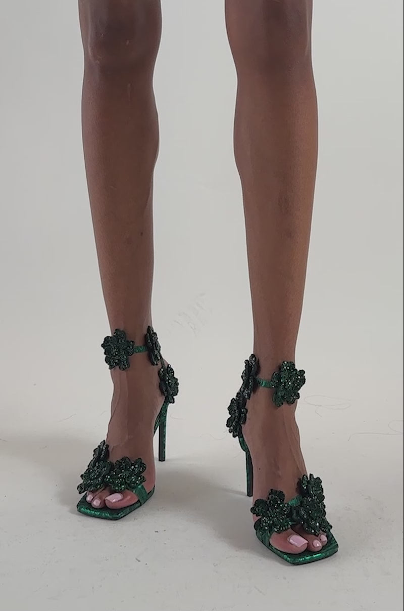Video of a woman modeling the AZALEA WANG DEBUTANTE GREEN RHINESTONE STILETTO SANDAL—green high-heeled sandals adorned with large floral appliqués and sparkling rhinestone accents that add a striking touch of glamor.