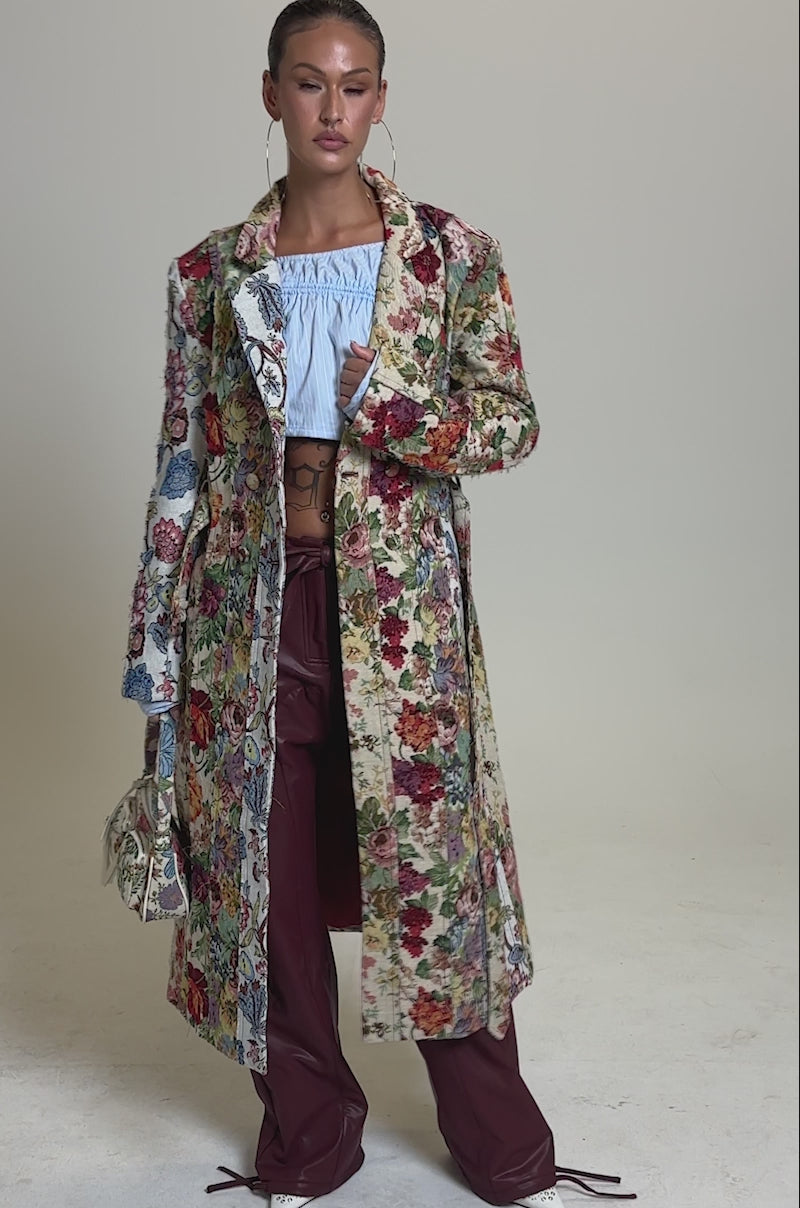 A video of a model wearing the SUNDAY PATCHWORK TRENCH over a light blue off-shoulder crop top and loose maroon pants, a confident woman holds a small handbag while posing against a plain background.