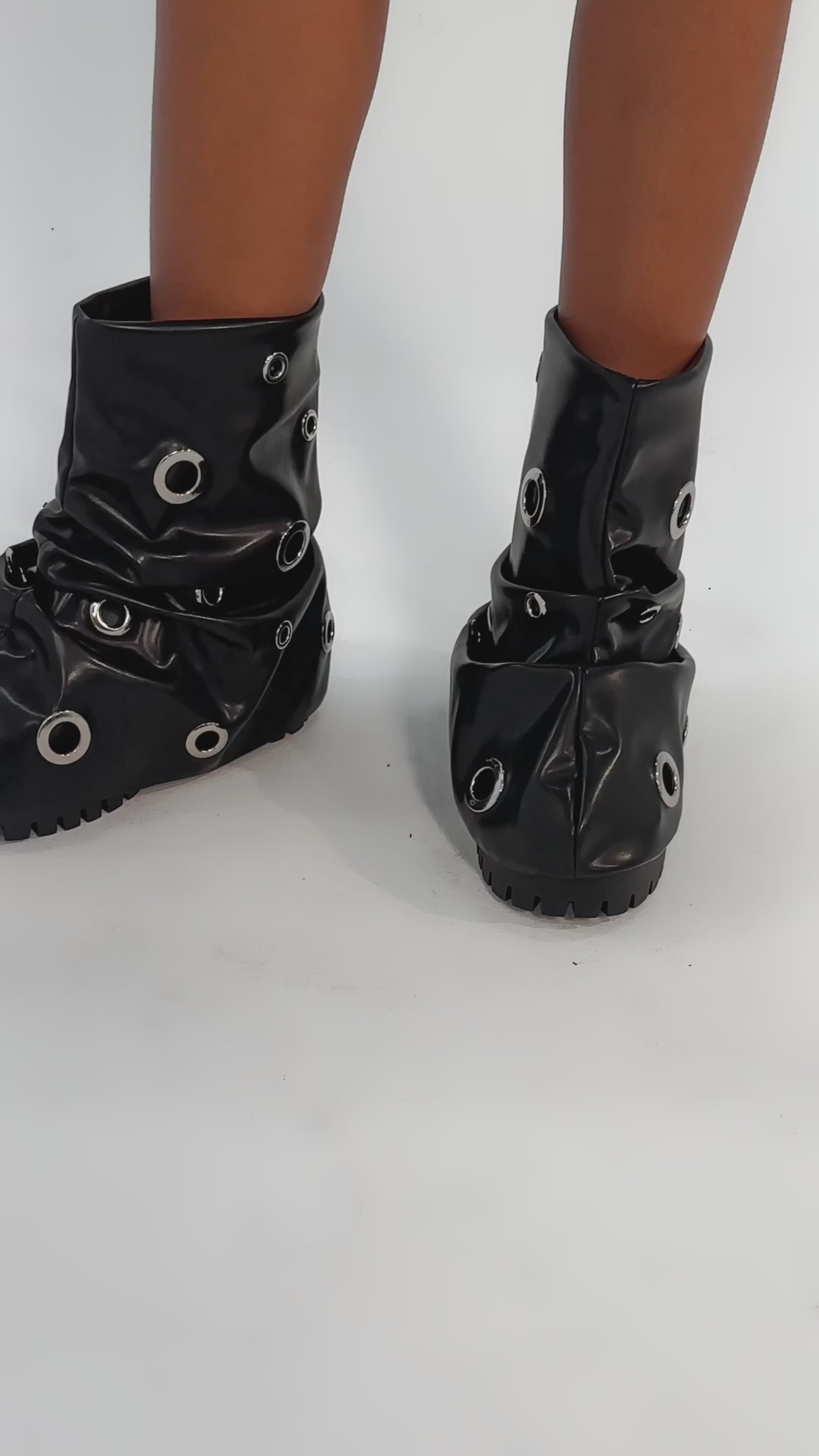 Video of woman modeling the AKIRA GOLD FABLE BLACK GROMMET EMBELLISHED FLAT BOOTIE with thick soles and large silver grommet holes, set against a light gray background.