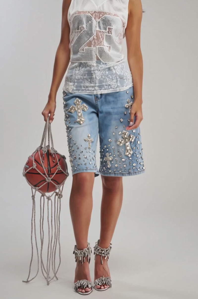 Video of woman modeling the BLESSED RHINESTONE BERMUDA, featuring knee-length denim embellished with large silver jeweled crosses and studs, styled with silver fringed high-heeled sandals.