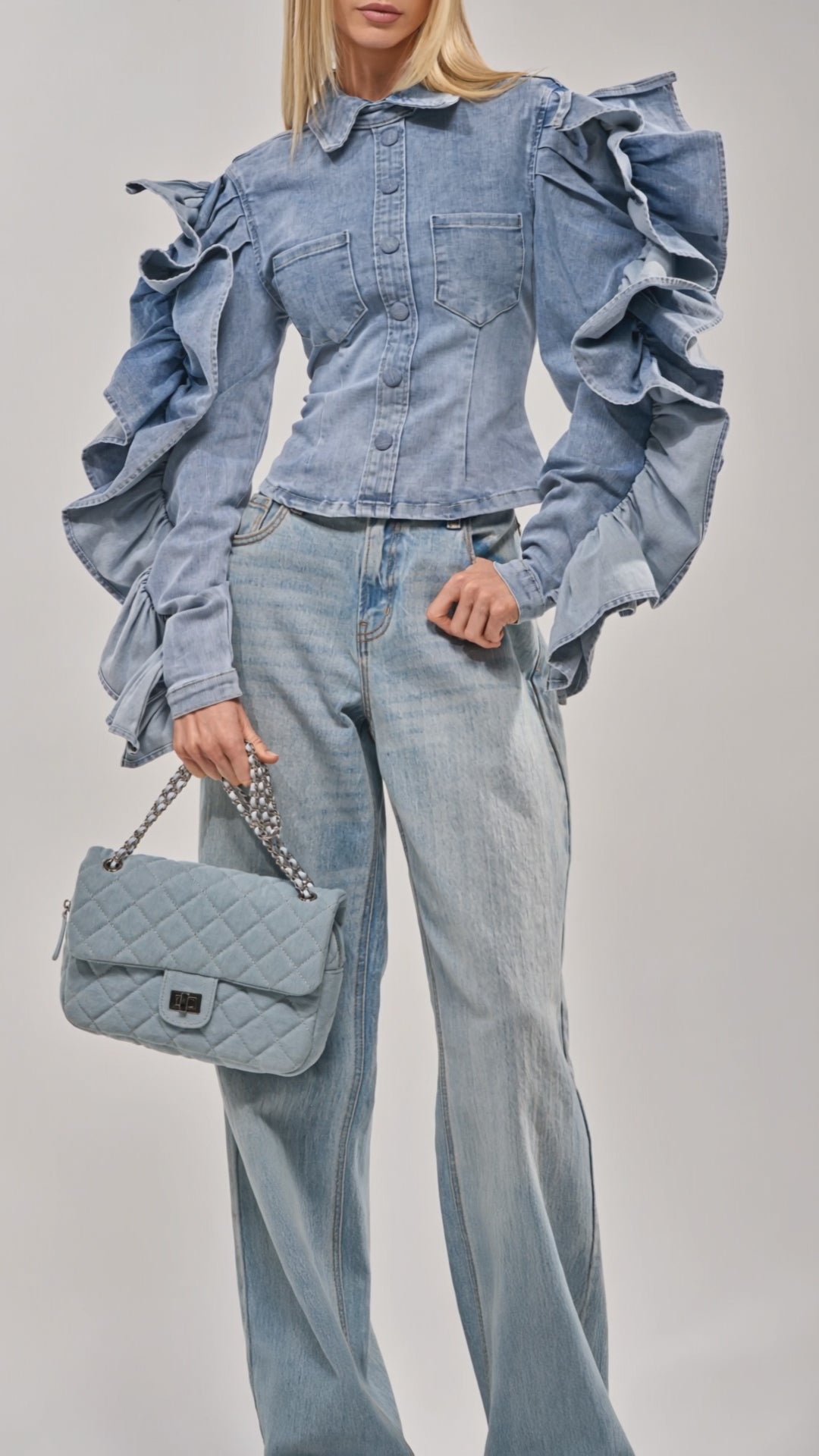 Video of woman modeling the DREA RUFFLE SLEEVE DENIM BUTTON DOWN with matching light blue jeans and a small quilted handbag. She has straight blonde hair and poses confidently against a plain background.