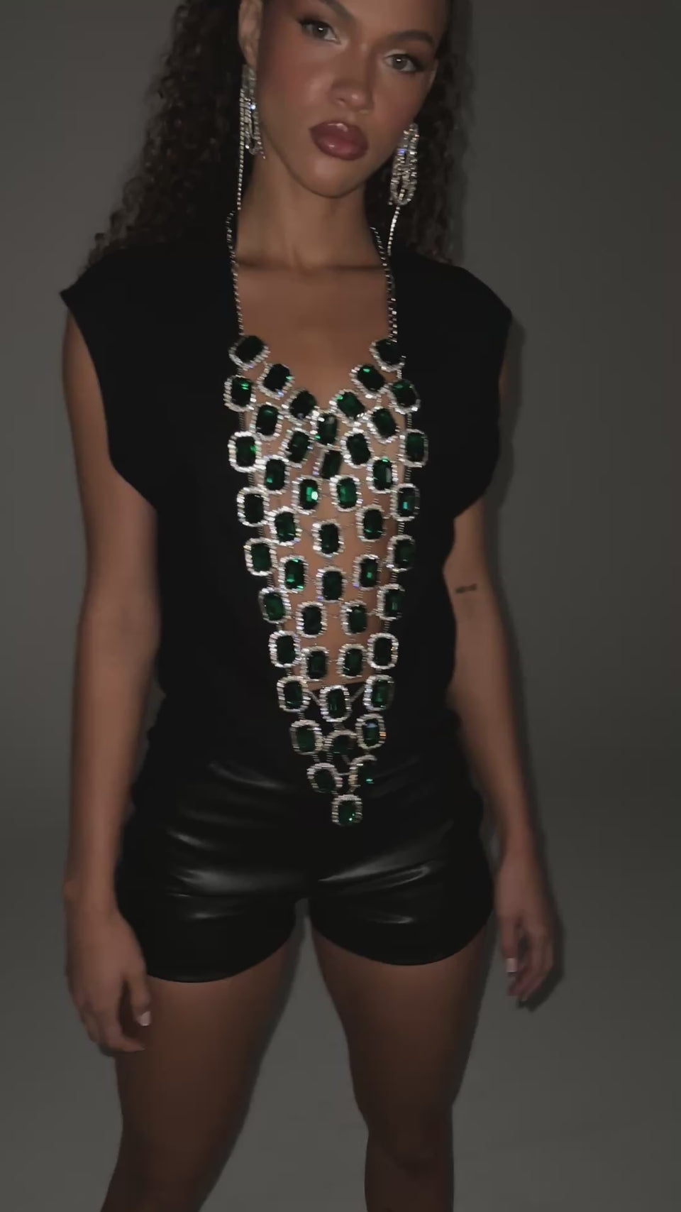 A video of a model wearing the EMERALD BABY RHINESTONE TEE. Crafted from a black stretch cotton blend fabric, this shimmering tee features a scoop neckline, cropped, sleeveless silhouette, and a front lattice panel of emerald rhinestone embellishments surrounded in a row of crystals and attached by chains with dangling halter straps with an adjustable lobster-clasp closure.