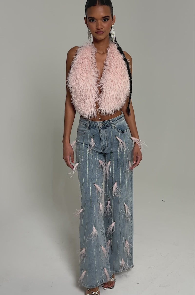 Video of woman modeling the ACT UP FUR BLOUSE, a fluffy light pink faux fur top, paired with dangling earrings and light blue jeans. She stands against a plain light background, looking confidently at the camera.