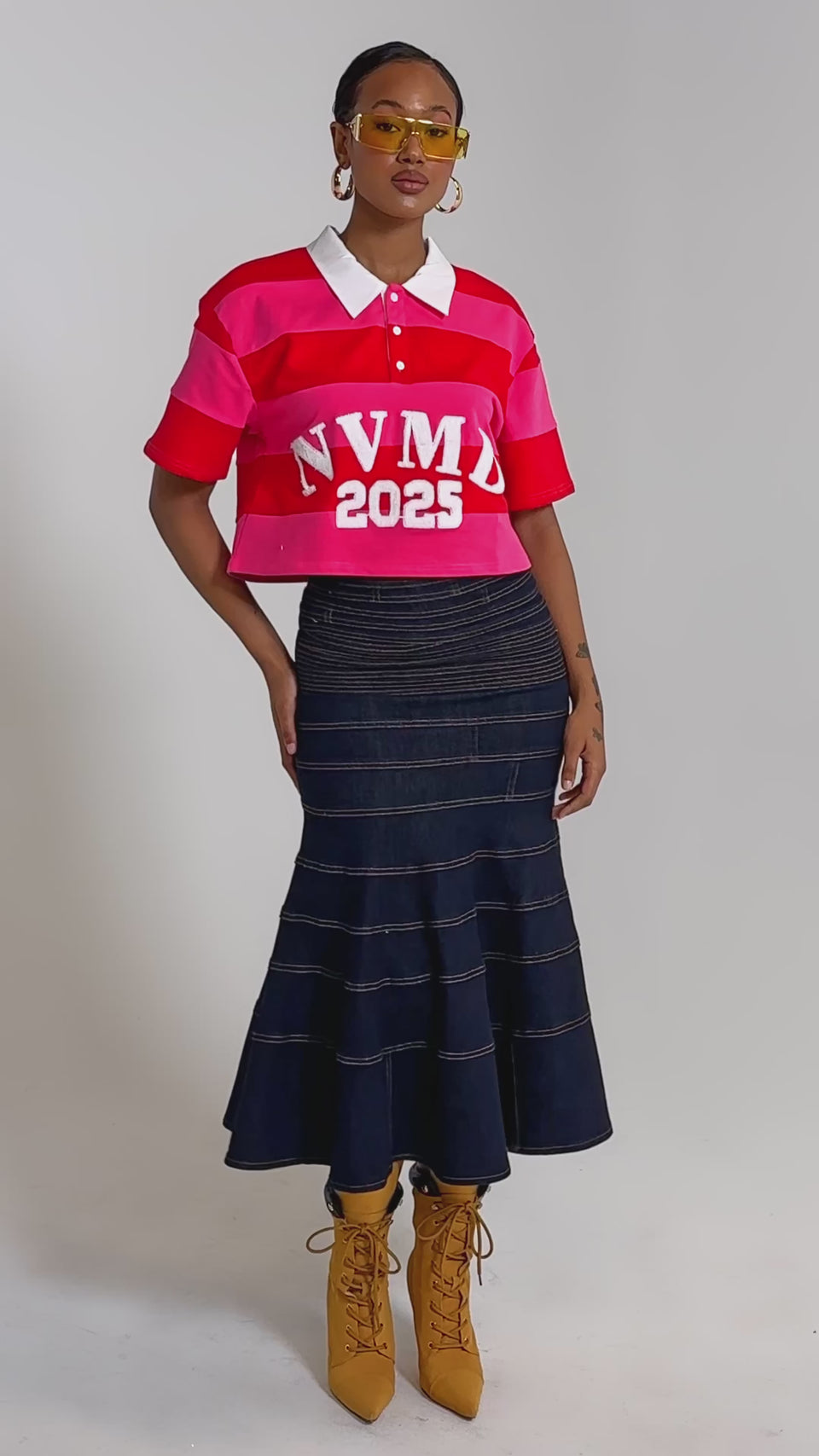 A video of a person in the NEVERMIND KNIT TEE with pink and red stripes reading "NVMD 2025," styled with a dark denim skirt, yellow heeled boots, and yellow-tinted sunglasses.