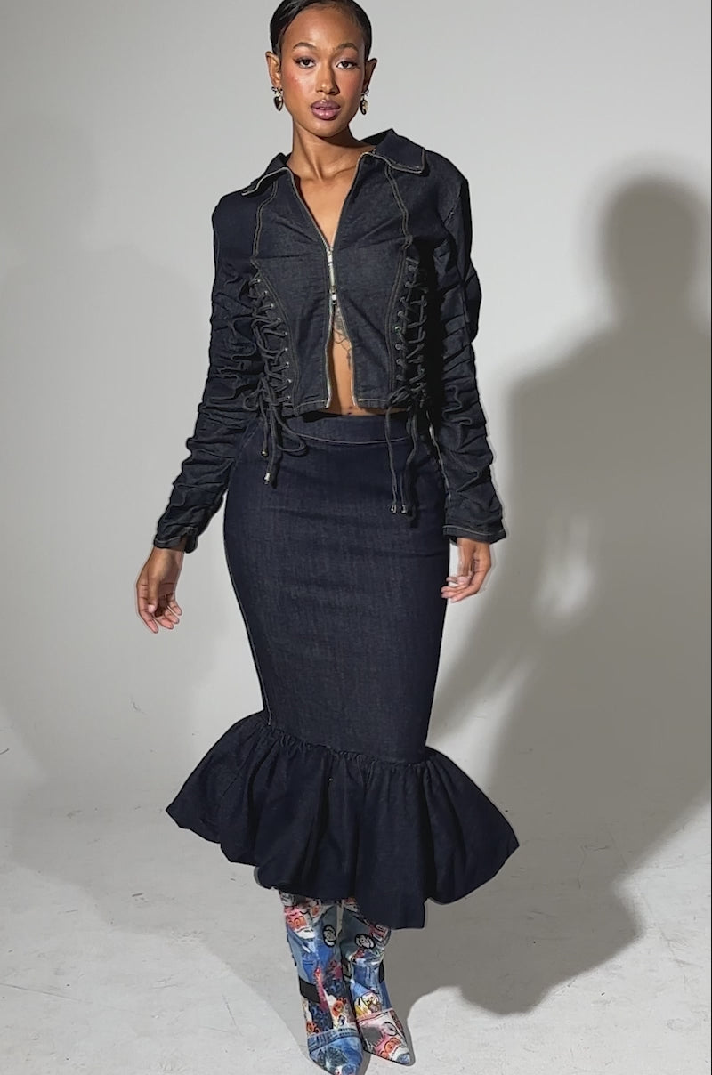 A video of a woman modeling the STRICTLY BUSINESS DENIM BLOUSE, a dark-wash jacket with ruched sleeves, paired with a matching flared mermaid skirt, colorful patterned knee-high boots, and hoop earrings against a plain light background.
