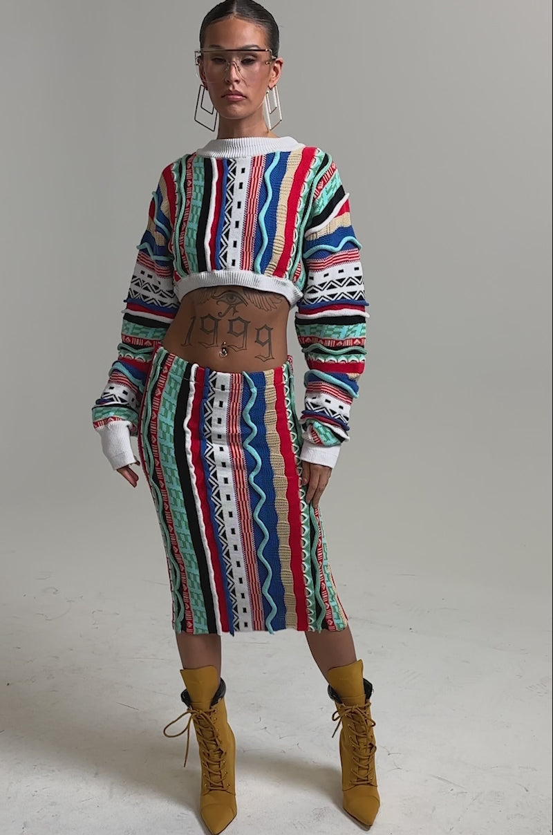 Video of woman modeling the FEELING THE VIBES CROP SWEATER and matching skirt, accessorized with geometric glasses, hoop earrings, and a “1999” stomach tattoo, set against a plain white background.