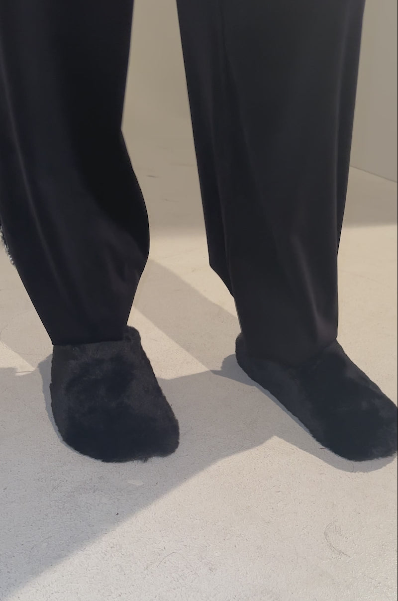 A video of a person wearing the AKIRA RHYS BLACK FUZZY FLAT and loose black pants with jeweled trim on one side, standing on a light surface. Only the lower legs and feet are visible.