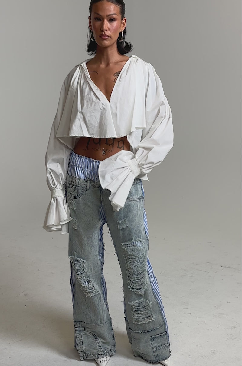 A video of a woman in a white ruffled crop top and the CALL IT WHAT YOU WANT MIXED UP TROUSER—high-waisted, wide-leg jeans with distressed patches and blue-and-white pinstripes—paired with hoop earrings and pointed white shoes.