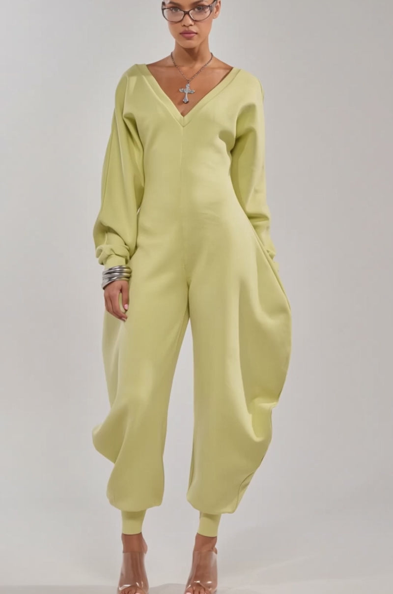 A video of a model wearing the COMFY COZY SWEATSHIRT JUMPSUIT IN LIME, featuring a contemporary streetwear look with voluminous legs and sleeves, styled with clear heels and silver jewelry.