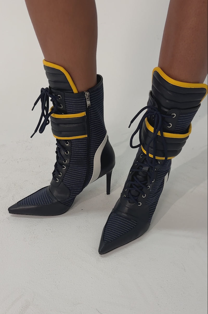 Video of woman modeling the AKIRA GOLD DEMAND NAVY STILETTO BOOTIE, featuring pointed toes, high heels, lace-up design, and a blue and yellow color scheme with sporty details, standing on a light background.
