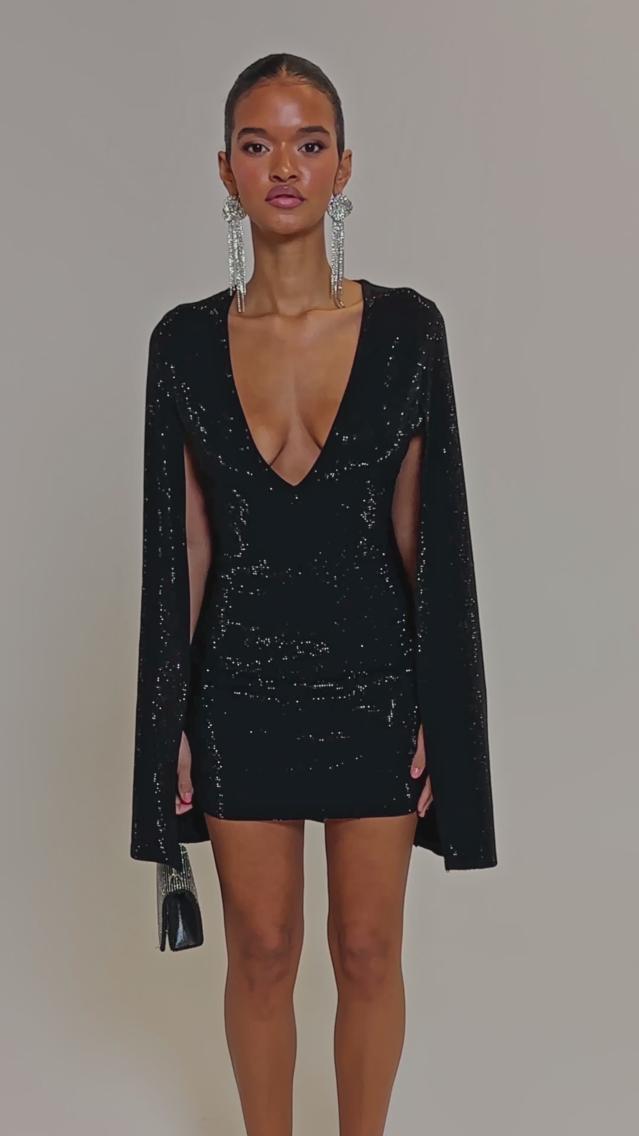 Video of woman modeling the LETS DANCE RHINESTONE MINI DRESS, featuring sparkling black rhinestones and cape sleeves, accessorized with long statement earrings and a small silver purse, stands against a plain light background.
