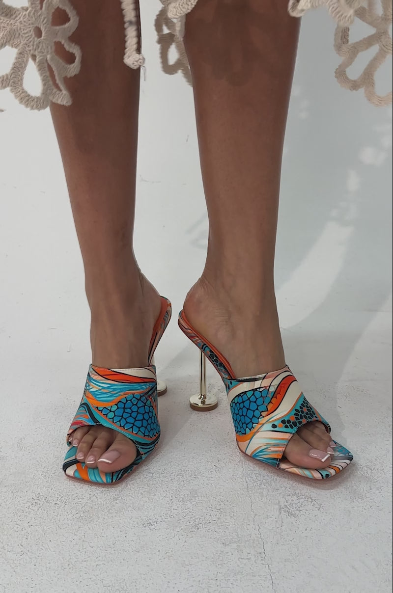 A video of a person wearing the AKIRA DOMNINA BLUE MULTI STILETTO PRINT SANDALS, featuring a bold blue, orange, and green pattern, unique metallic gold heels, and comfort cushion insole.