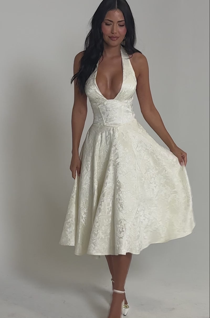 Video of woman modeling the SWEET CAROLINE HALTER MIDI DRESS—a cream-colored corset halter style with a deep V-neckline and flared mid-calf skirt, perfect for bridal events—finished with white high heels.