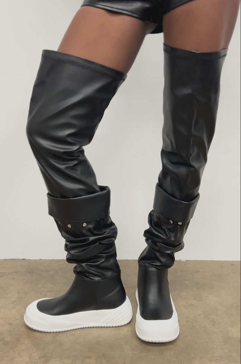 Video of woman modeling the AZALEA WANG SMARTER BLACK RUCHED PU FLAT BOOT with white rubber soles and black faux leather shorts stands against a white wall.