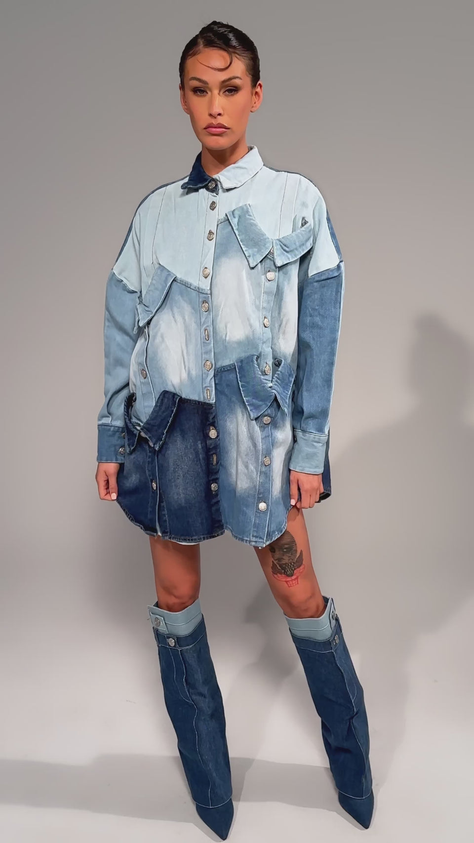 Video of woman modeling the MONDAY BLUES DENIM MINI DRESS with a relaxed patchwork silhouette, large buttons, flap pockets, and varying blue denim shades, a person with dark tied-back hair shows off an upper thigh tattoo.