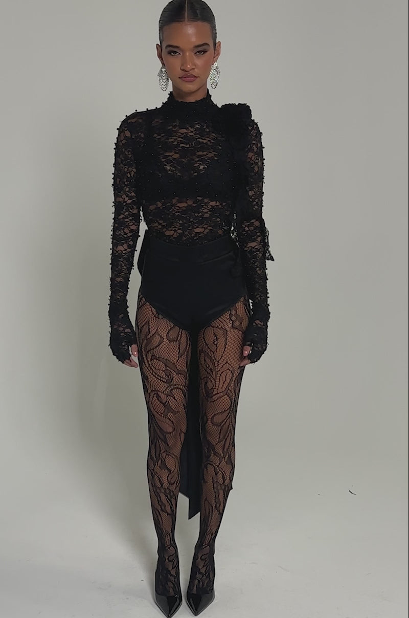 Video of woman modeling the LACED IN LOVE BLOUSE—a sheer floral lace, open-back design—she pairs it with black high-waisted bottoms and patterned tights, accessorizing with statement earrings and slicked-back hair for a confident pose.