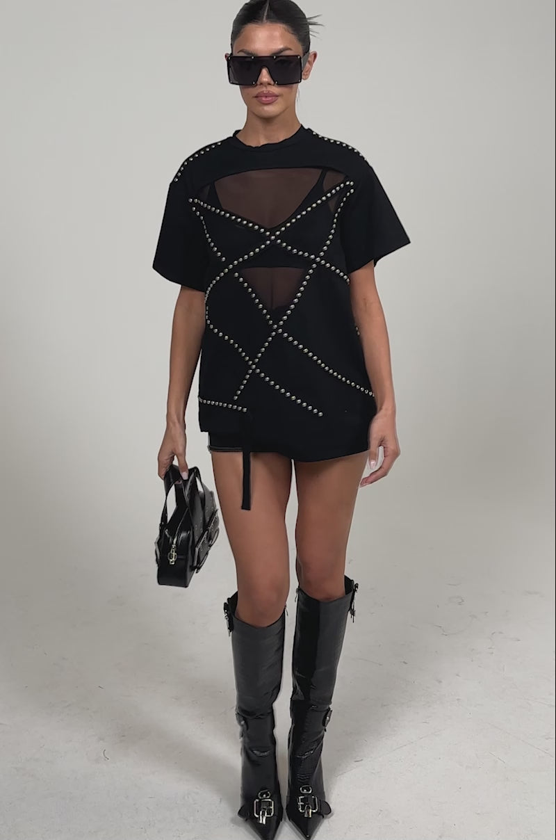 Video of woman modeling the MISTA RAJ OVERSIZED TEE with silver studs and mesh paneling stands against a plain background, holding a black bag.