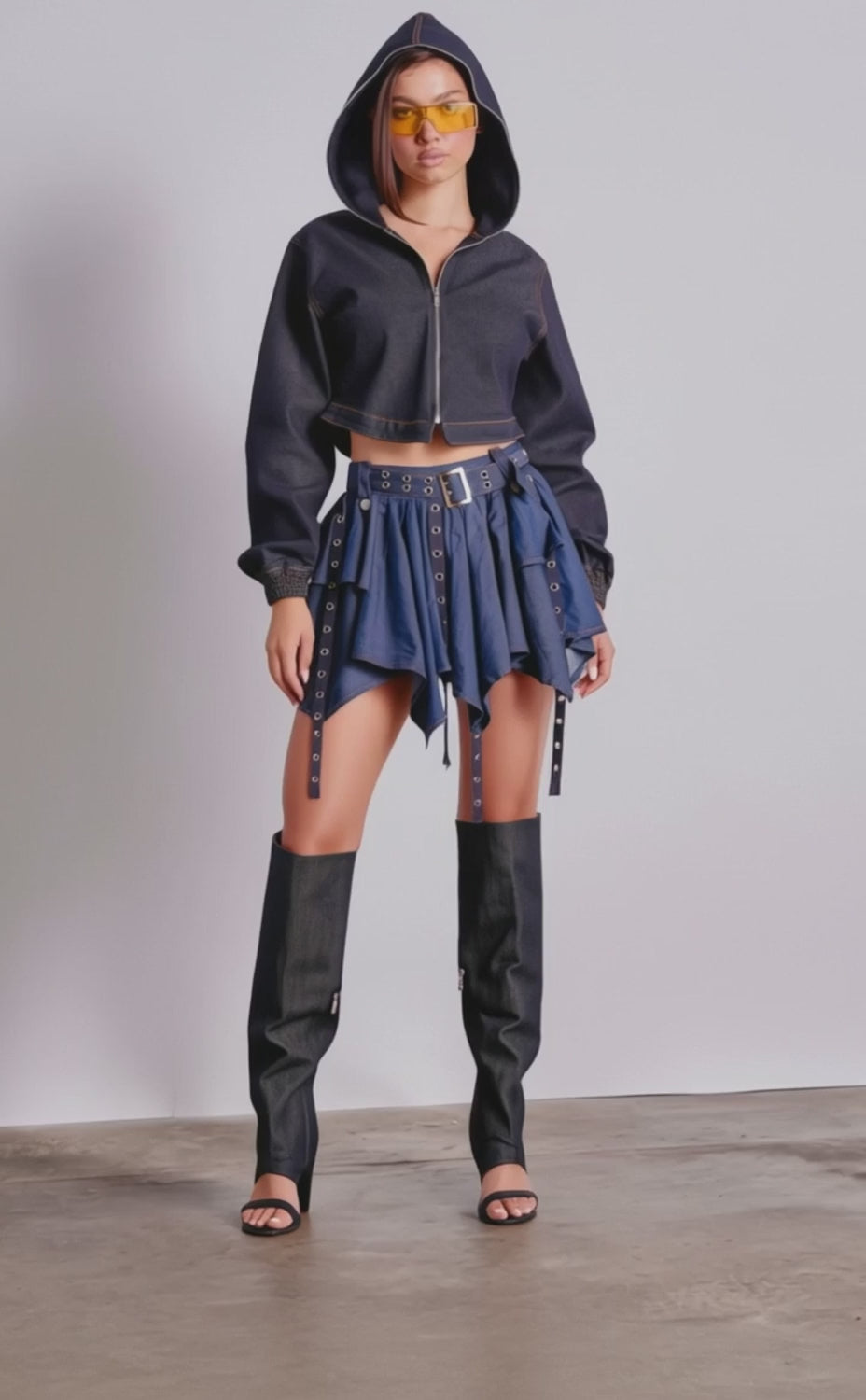 A video of a woman wearing the BEAM ME UP HOODED DENIM JACKET, paired with a blue asymmetrical mini skirt, tall black denim leg warmers, black heels, and yellow sunglasses.
