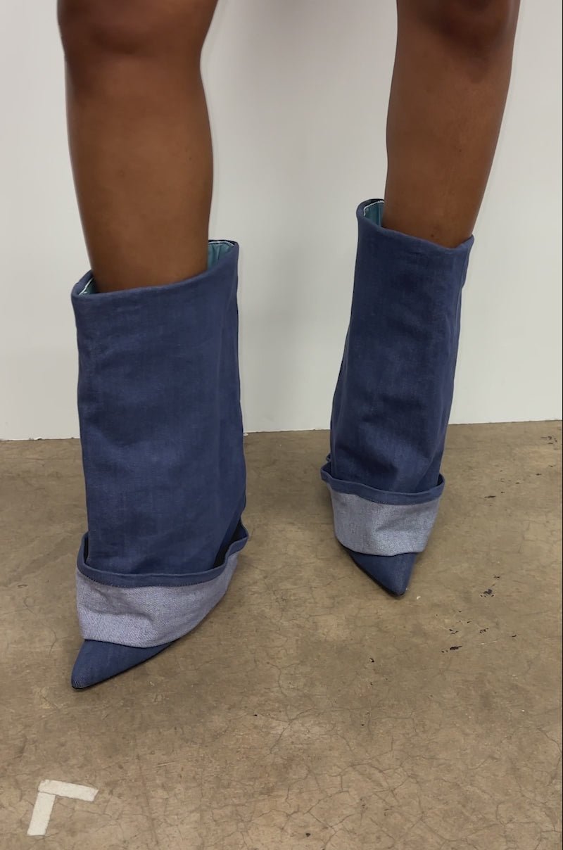 A video of a person wearing the AZALEA WANG JATIN DENIM CUFF FOLD OVER WEDGE BOOTIE, styled mid-calf and pointed toe, standing against a white wall on a concrete floor.